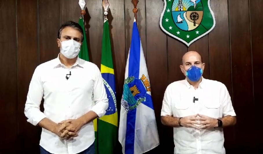 governador e prefeito lado a lado de máscara, posando para a câmera e com bandeiras ao fundo